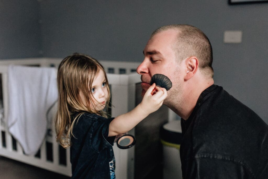 Young boy applying makeup with a brush to his dad. 